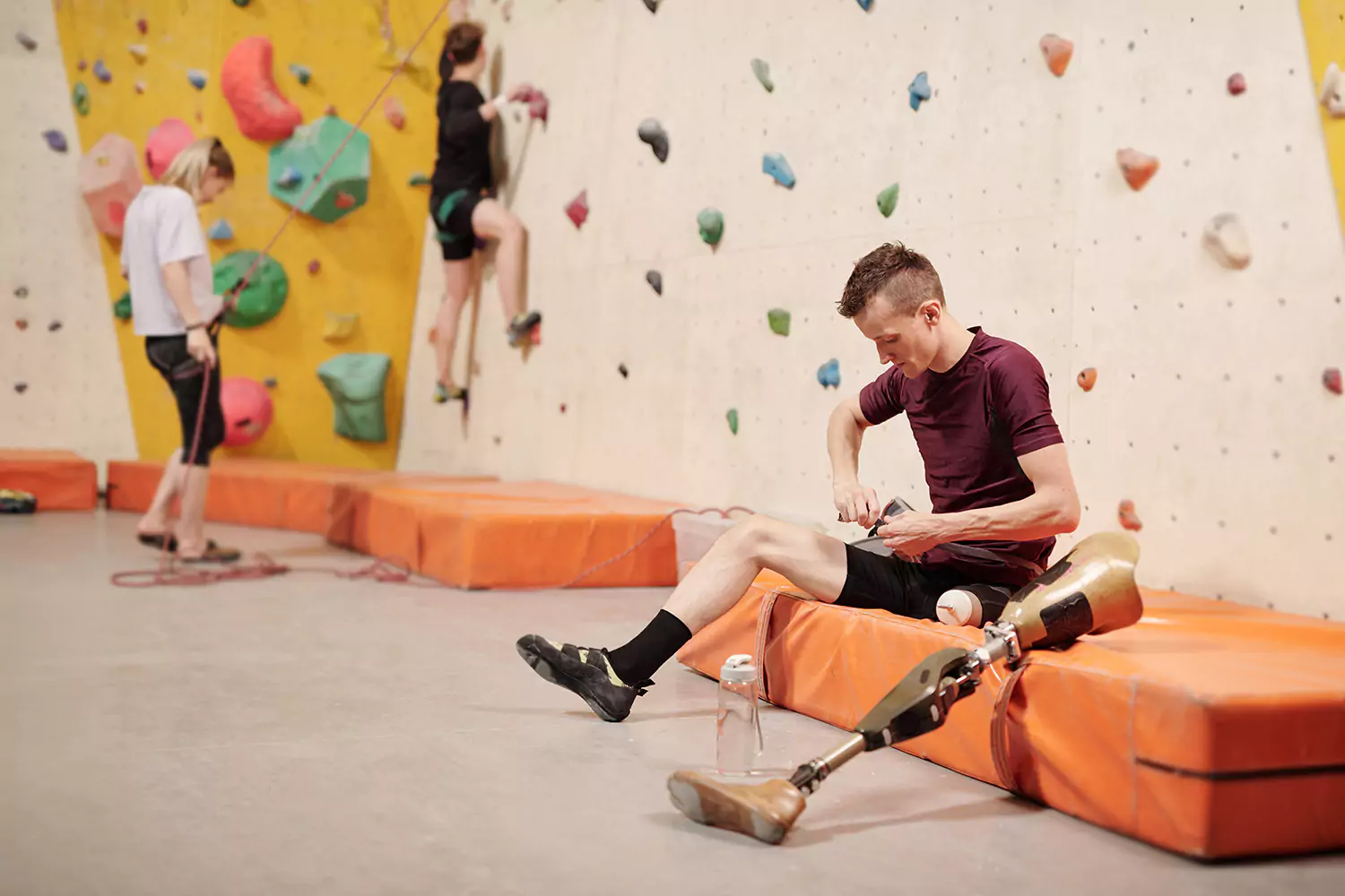 A young man with an amputated leg is sitting on a matt while at a rock climbing gym